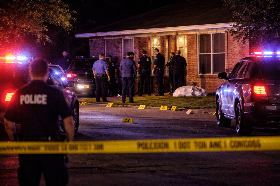 Nighttime police investigation outside a brick house with officers, patrol cars, and yellow evidence markers on the lawn.