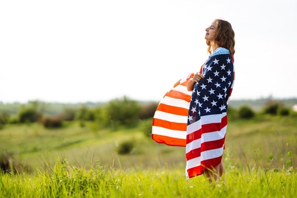 Woman draped in an American flag stands in a green field, looking up with a serene expression (patriotism/image of national pride).