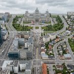 Aerial view of a city: a grand neoclassical capitol building with a dome, flagpoles, and a green plaza, surrounded by modern skyscrapers and streets.] ,