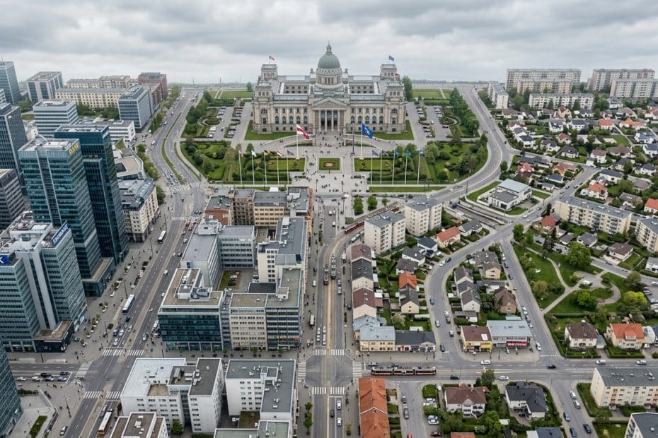 Aerial view of a city: a grand neoclassical capitol building with a dome, flagpoles, and a green plaza, surrounded by modern skyscrapers and streets.] ,