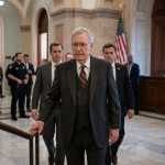 Older man in a dark pinstripe suit leads a group down a marble hallway, hand on a railing, with reporters and security nearby and an American flag in the background.