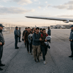 Group of travelers walking in a line on an airport tarmac, flanked by security guards near a large airplane.